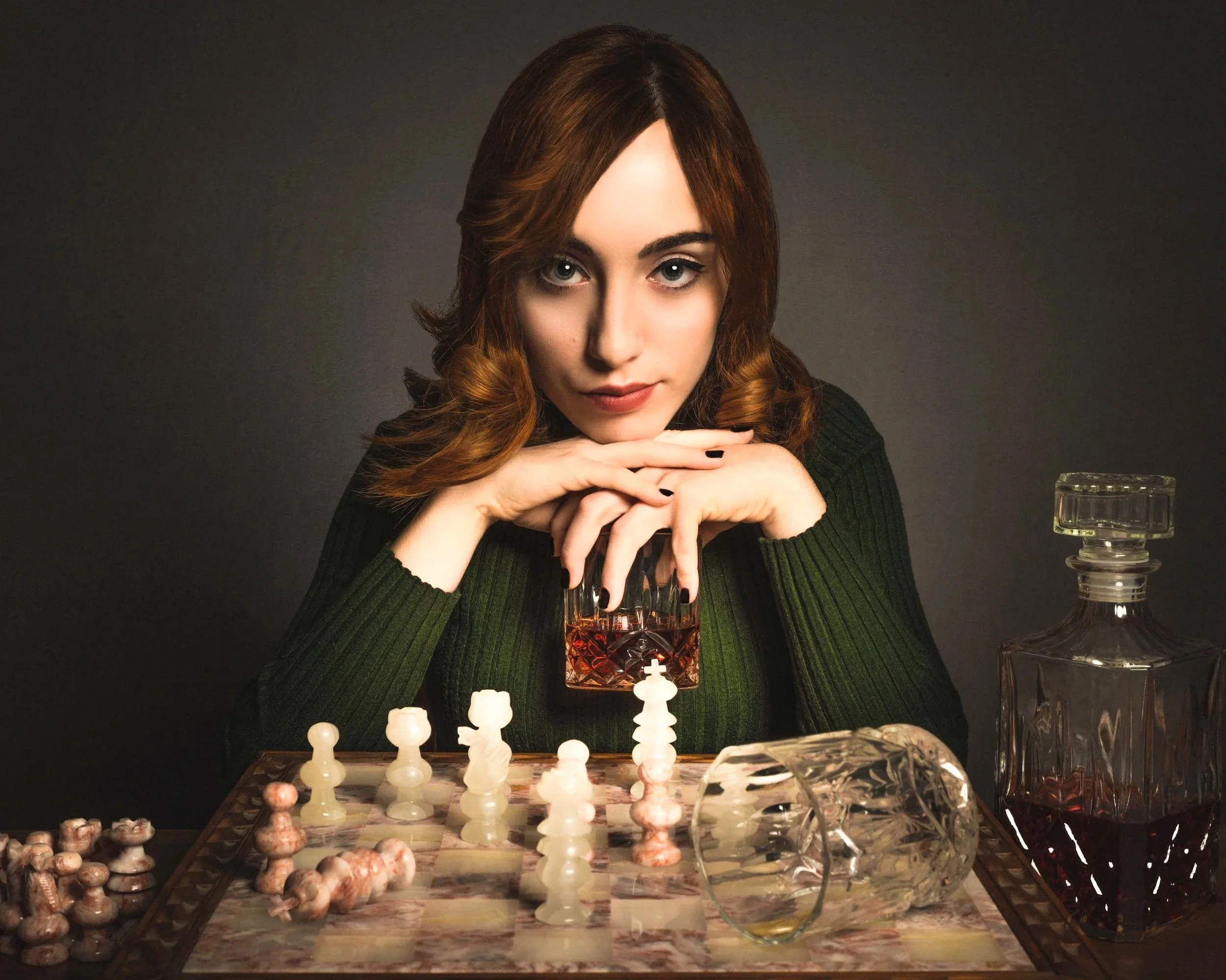 A woman with brown hair and hazel eyes sitting at a table playing chess, surrounded by glass decorative objects and a glass of water, against a dark background.