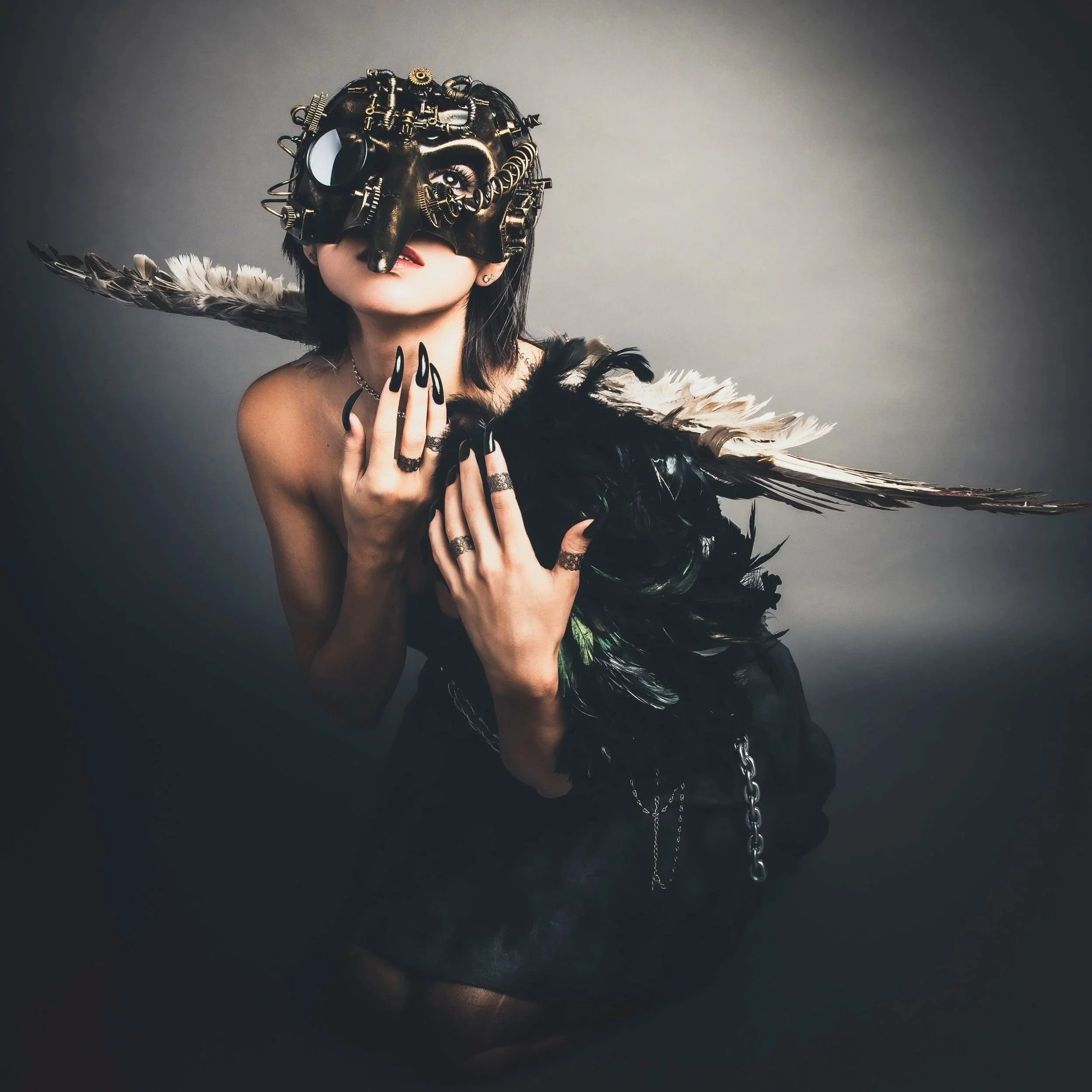 A person wearing a black mask with metallic details and eclectic accessories, holding feathers, against a dark background.