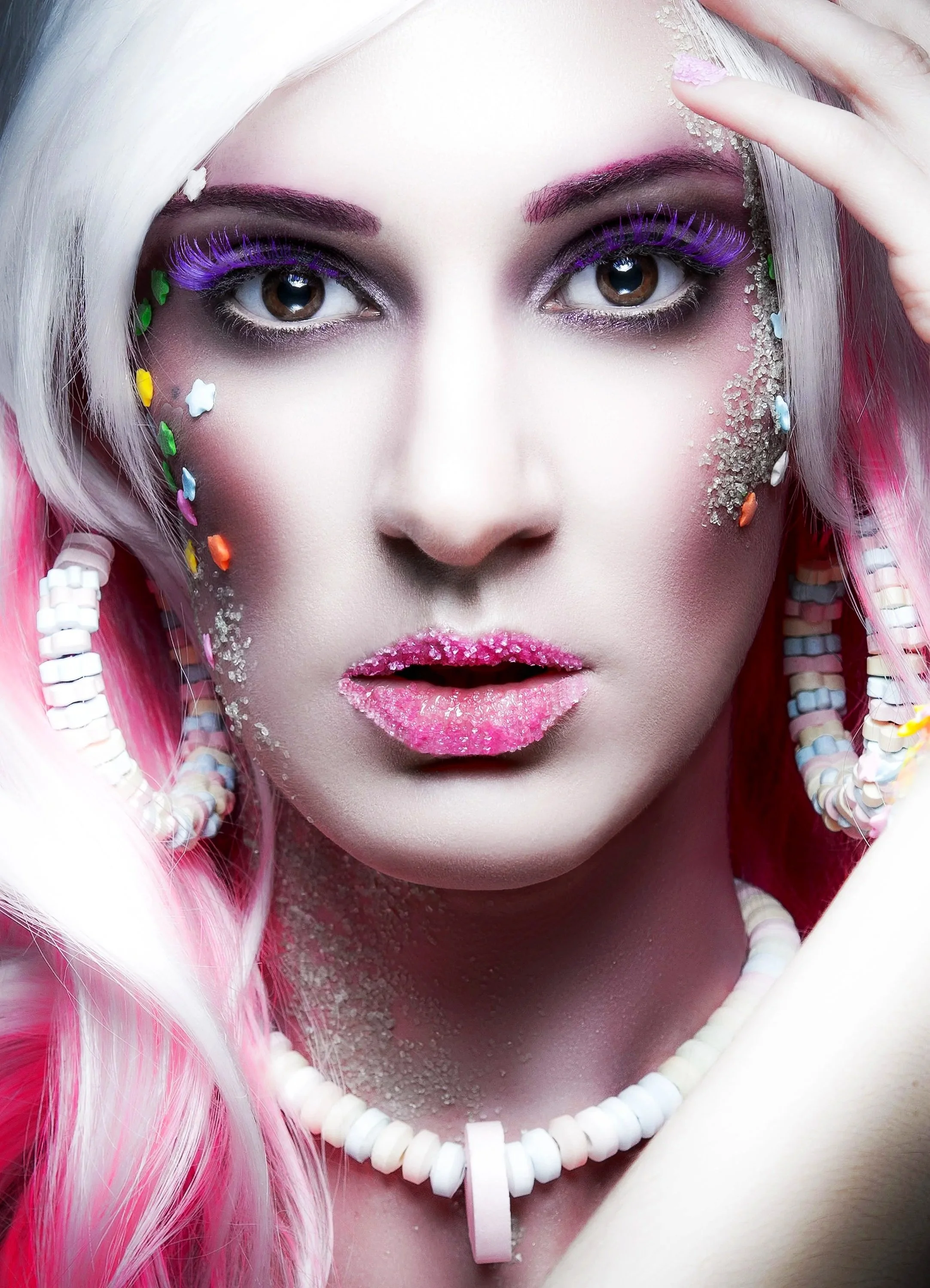 Close-up of a woman with vibrant makeup, pink hair, and colorful beads in her hair. She is wearing purple eyelashes, pink lipstick with glitter, and has multicolored small decorative beads on her face. She also wears a white beaded necklace and holds