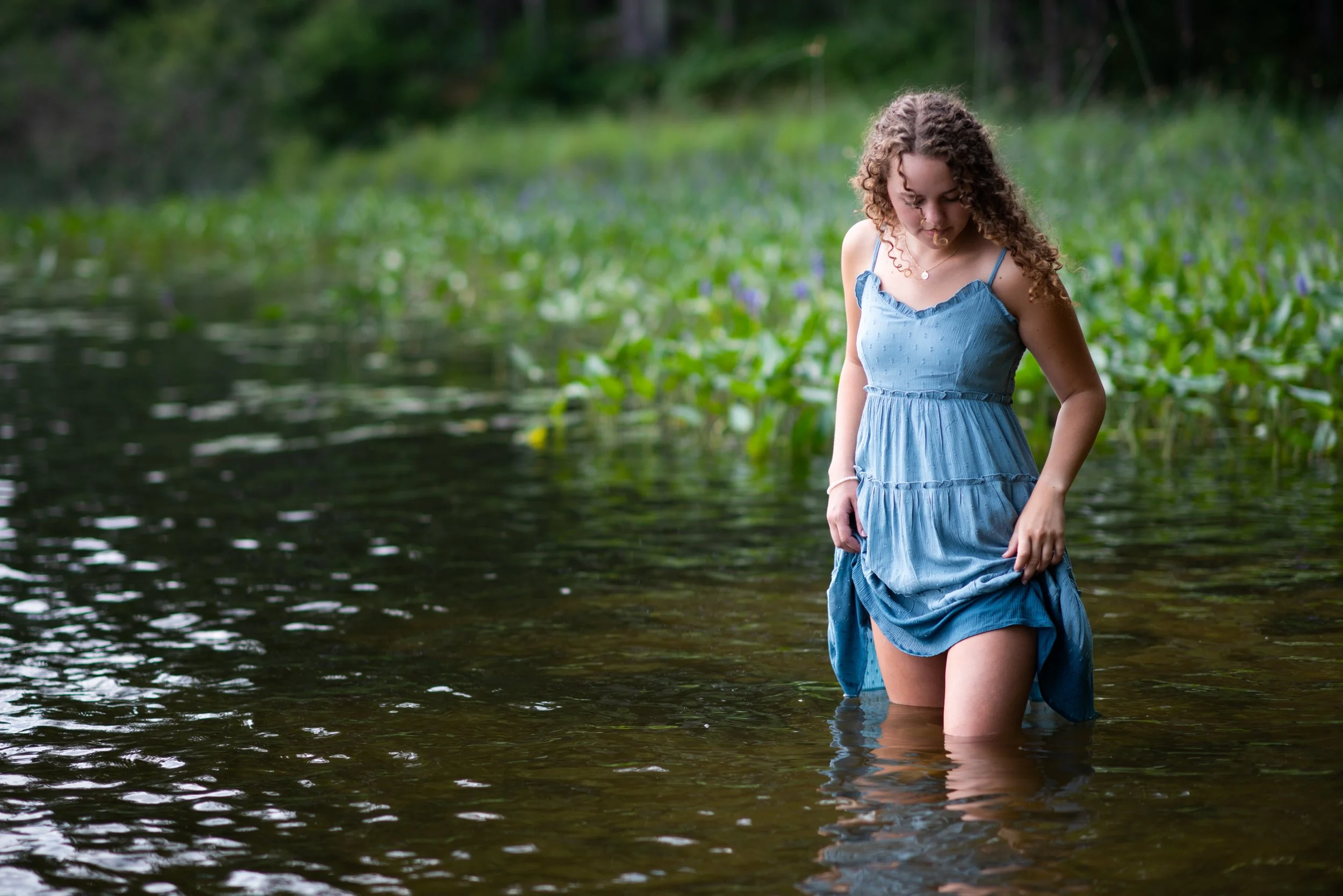 A girl with curly brown hair in a blue dress standing knee-deep in a calm body of water surrounded by lush greenery.