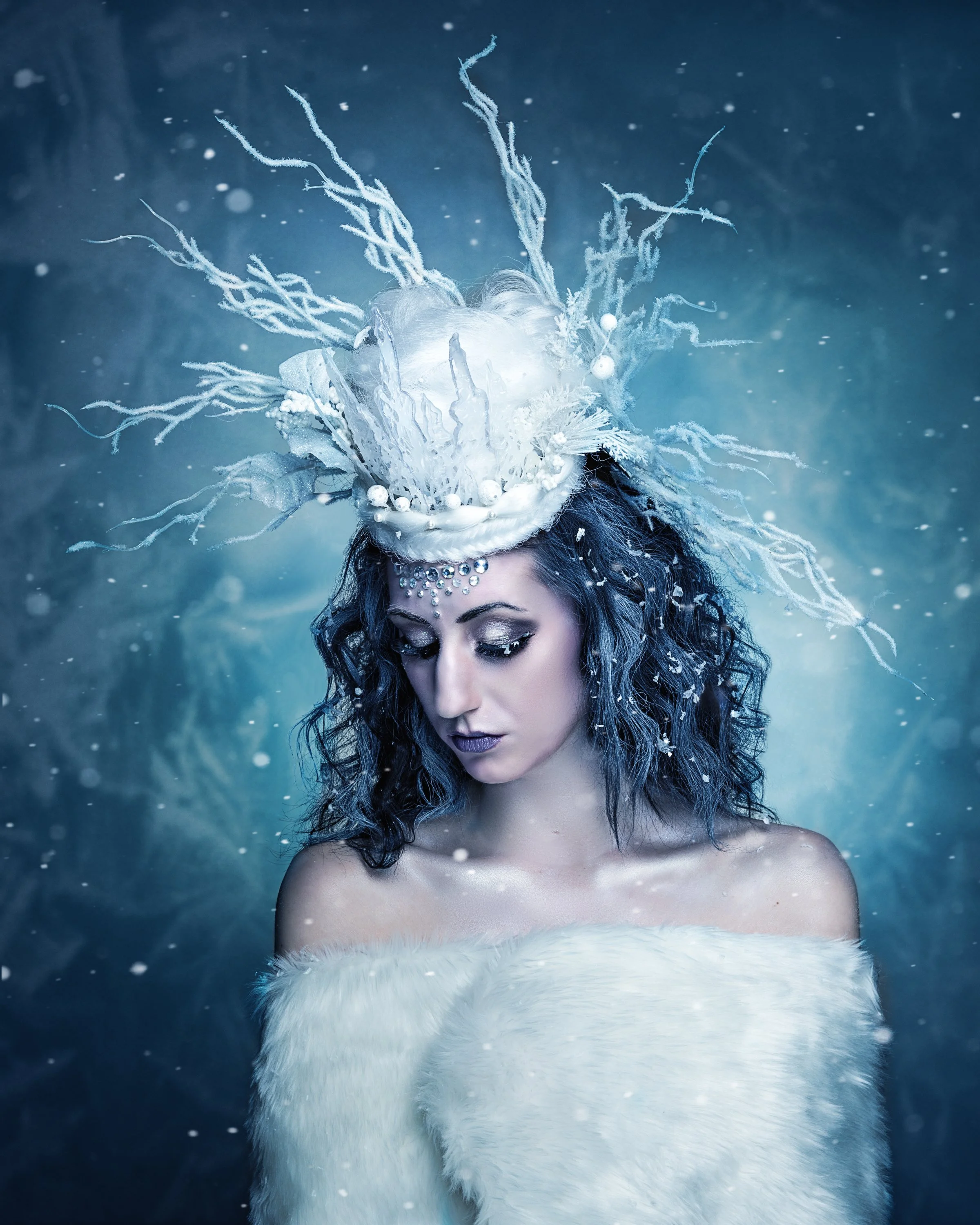 A woman with dark curly hair and makeup, wearing a white fur garment, with a detailed elaborate headpiece made of branches, white ornaments, and decorative elements, against a snowy, icy blue background.
