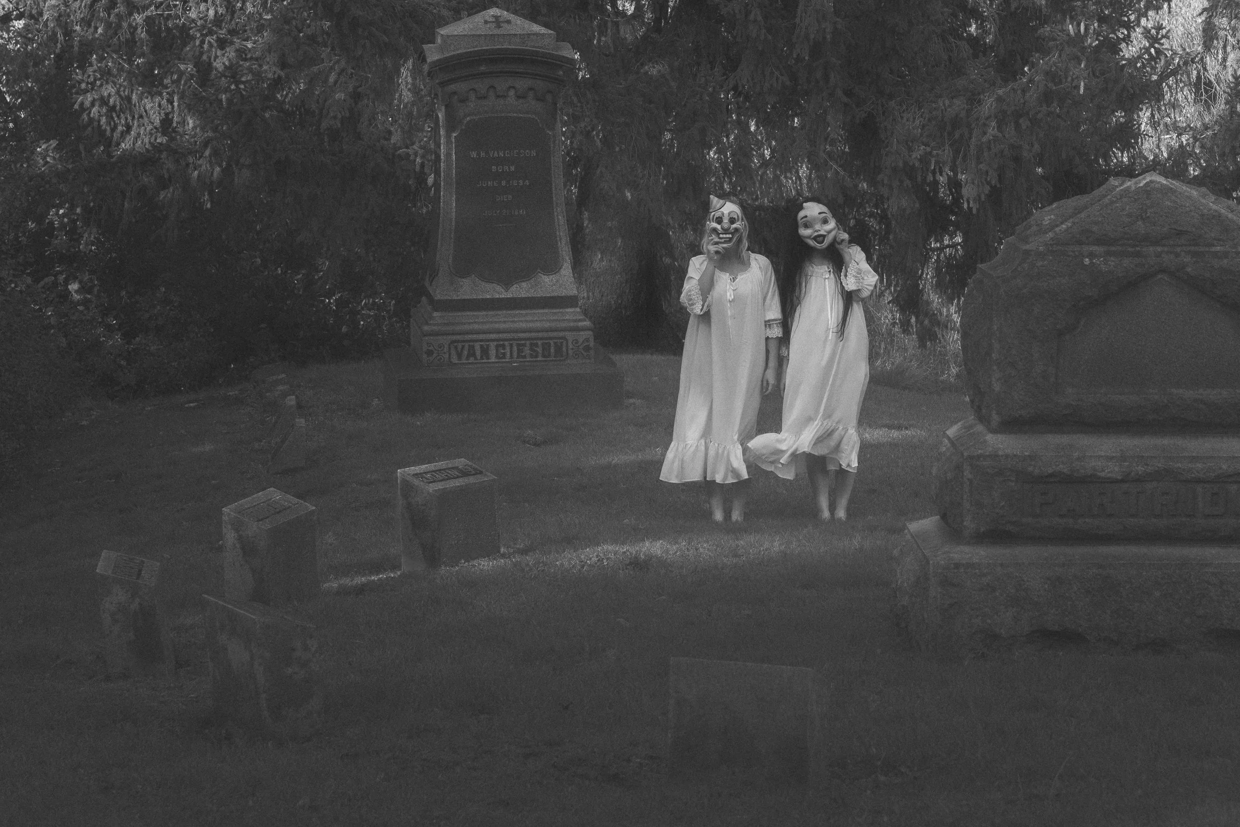 Two women in white dresses standing in a graveyard, wearing masks that resemble a clown and a cartoon character, with trees and gravestones behind them.