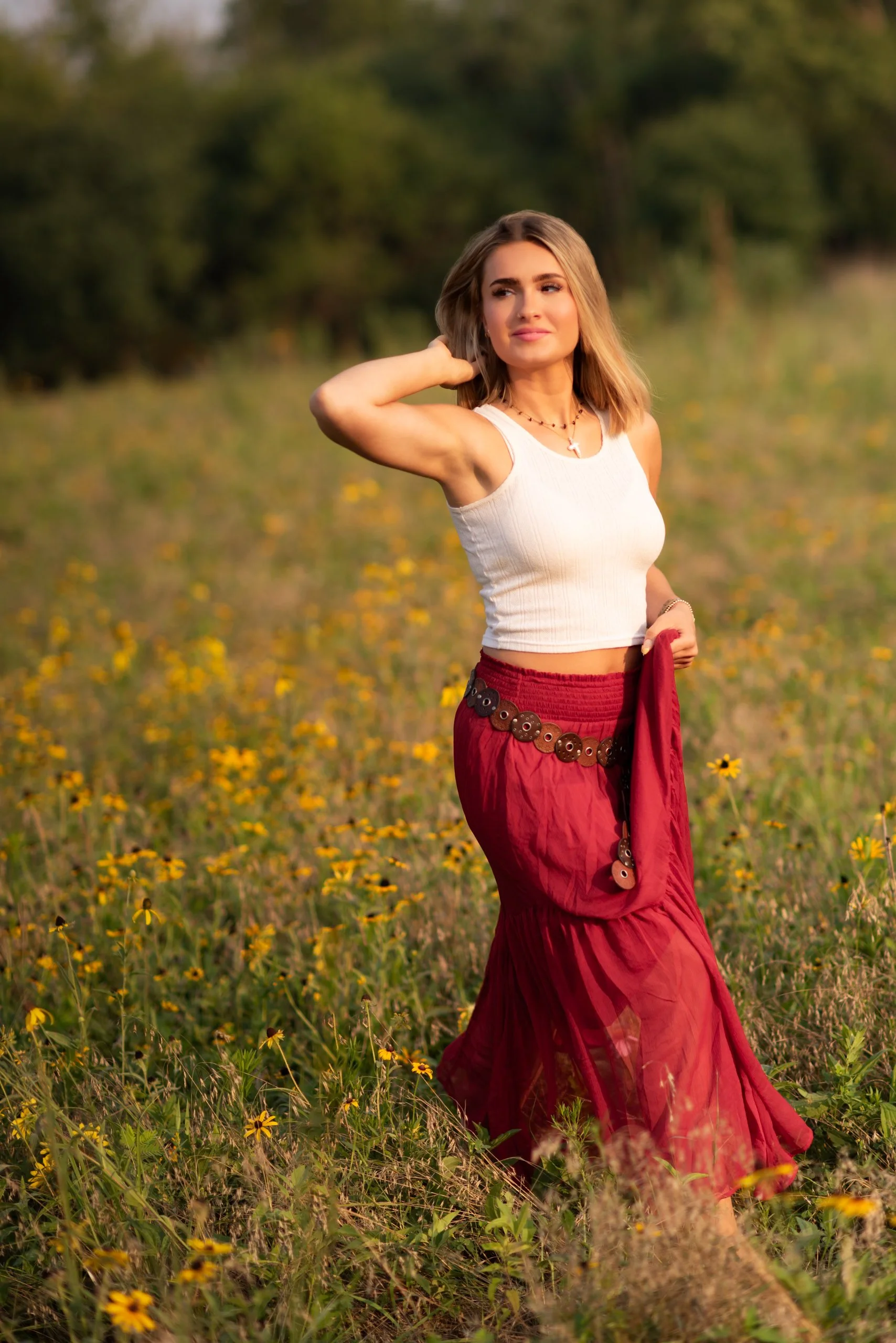 A young woman with short blonde hair standing in a field of yellow flowers during sunset. She is wearing a white sleeveless crop top and a red skirt with a wooden belt, and she has a cross necklace. She is touching her hair with her right hand and holding a red cloth in her left hand, with a serene expression on her face.