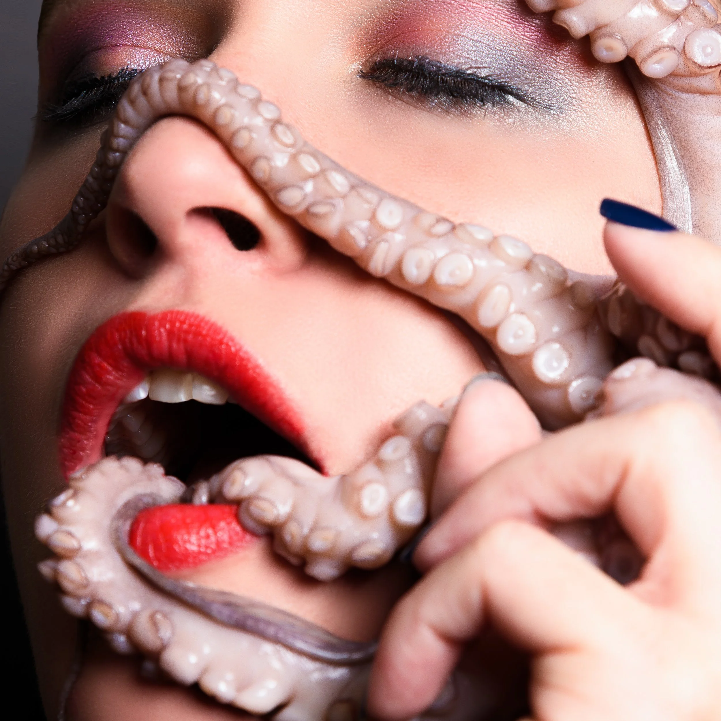 Close-up of a person's face with colorful makeup, red lipstick, closed eyes, and an octopus tentacle covering some of their face.