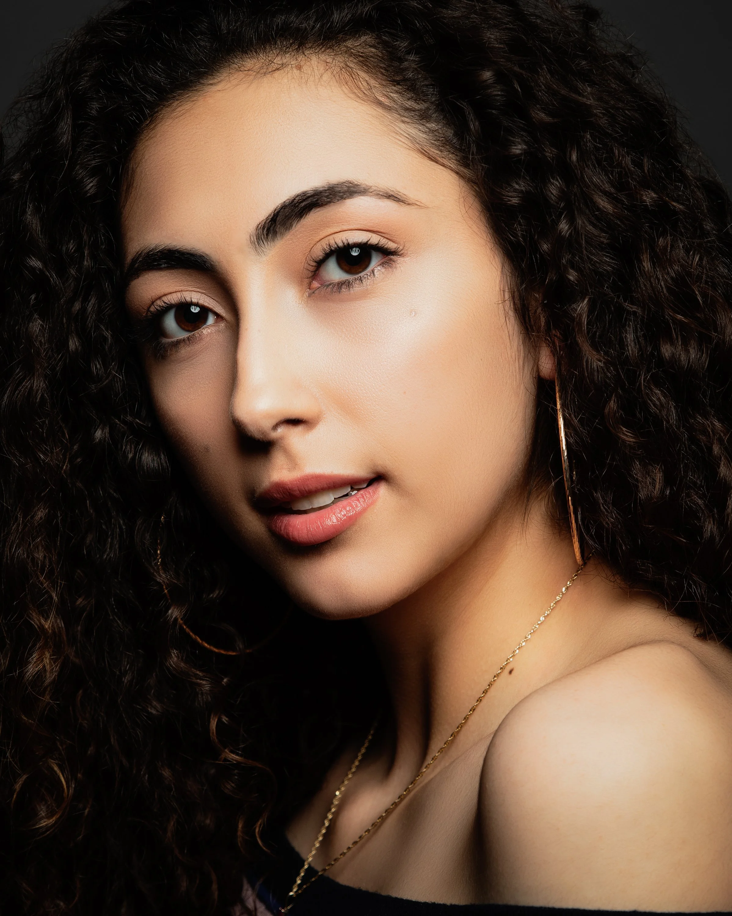 Close-up of a young woman with curly dark hair, wearing subtle makeup, gold necklaces, and a strapless black top, smiling softly in front of a dark background.