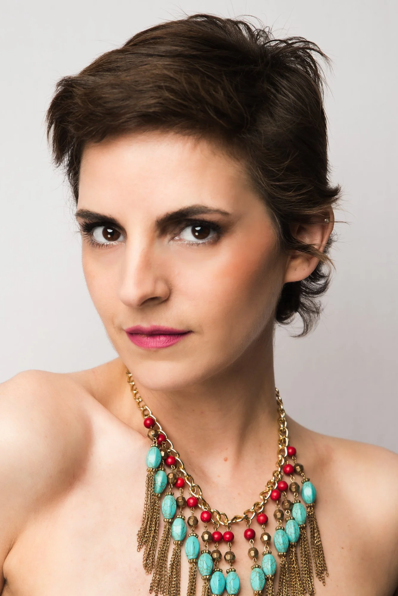A woman with short brown hair, dark eye makeup, pink lipstick, wearing a gold necklace with turquoise and red beads, posed with her right shoulder bare against a plain background.