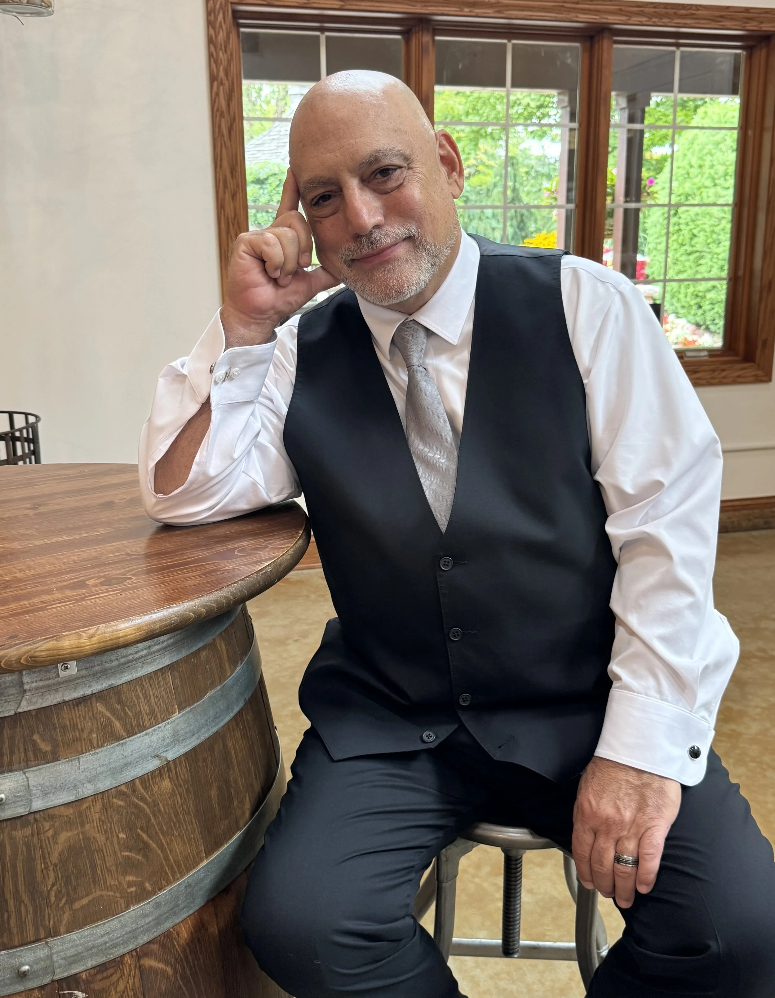 A bald man with a white beard and mustache, wearing a white dress shirt, gray tie, and black vest, sitting by a wooden bar table near large windows with wooden frames, with a green outdoor view in the background.
