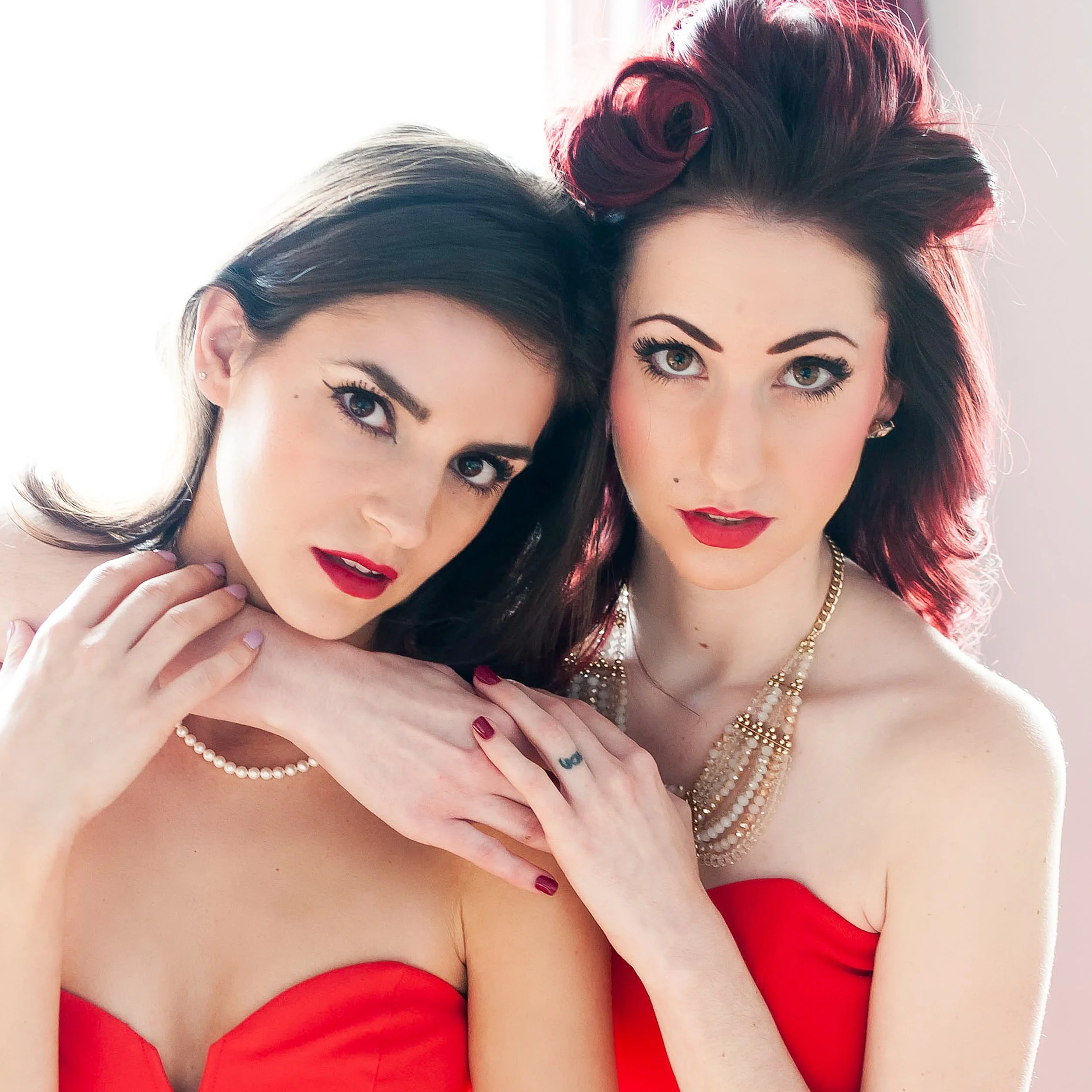 Close-up of two women wearing red dresses and jewelry, posing together, with visible faces, hair, and makeup.