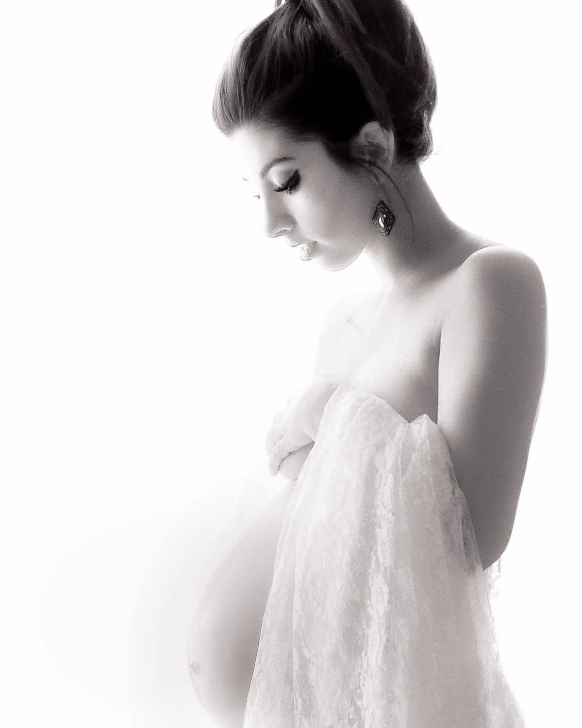 A black-and-white photograph of a pregnant woman with her eyes closed, wearing earrings, and holding a floral fabric close to her chest.