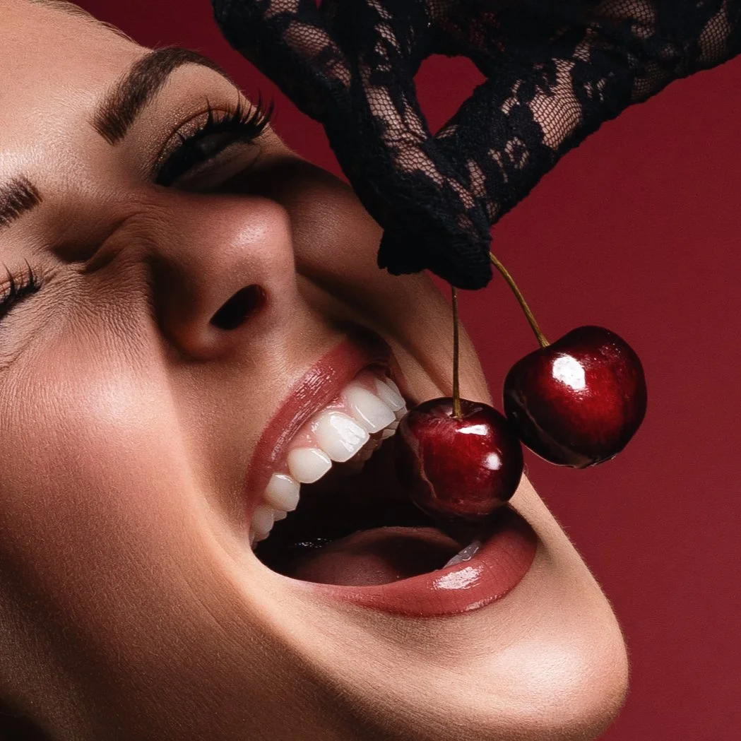 Close-up of a smiling woman with cherries held above her open mouth, wearing lace gloves, against a red background.