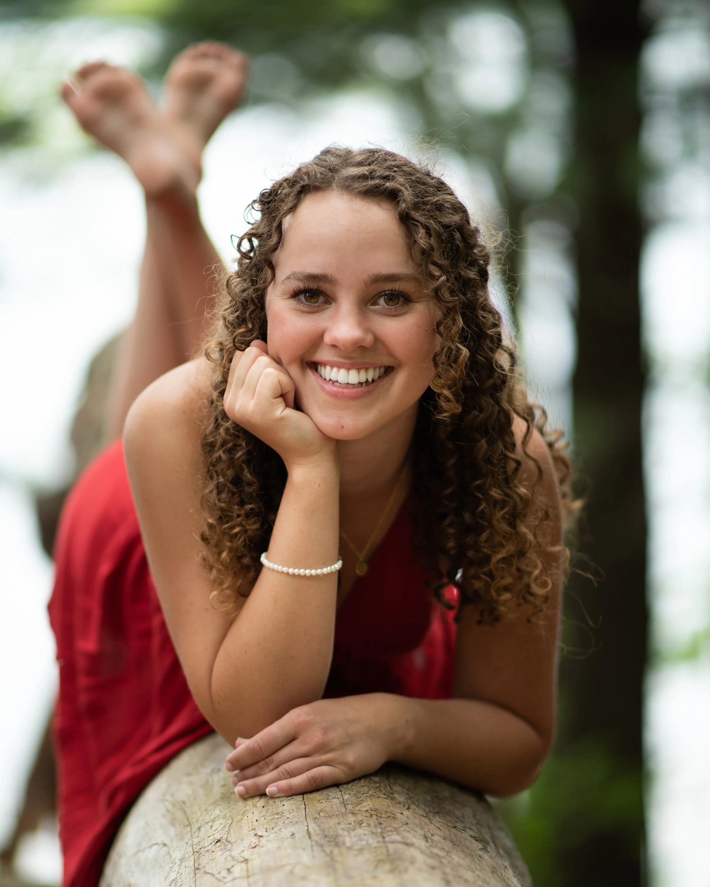 A smiling woman with curly hair lying on a fallen tree trunk outdoors, with her chin resting on her hand, in a lush green environment.