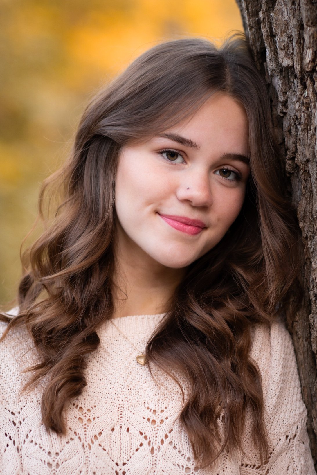 A young woman with long, wavy brown hair, wearing a beige knitted sweater, leaning against a tree and smiling softly, with a blurred autumn background.
