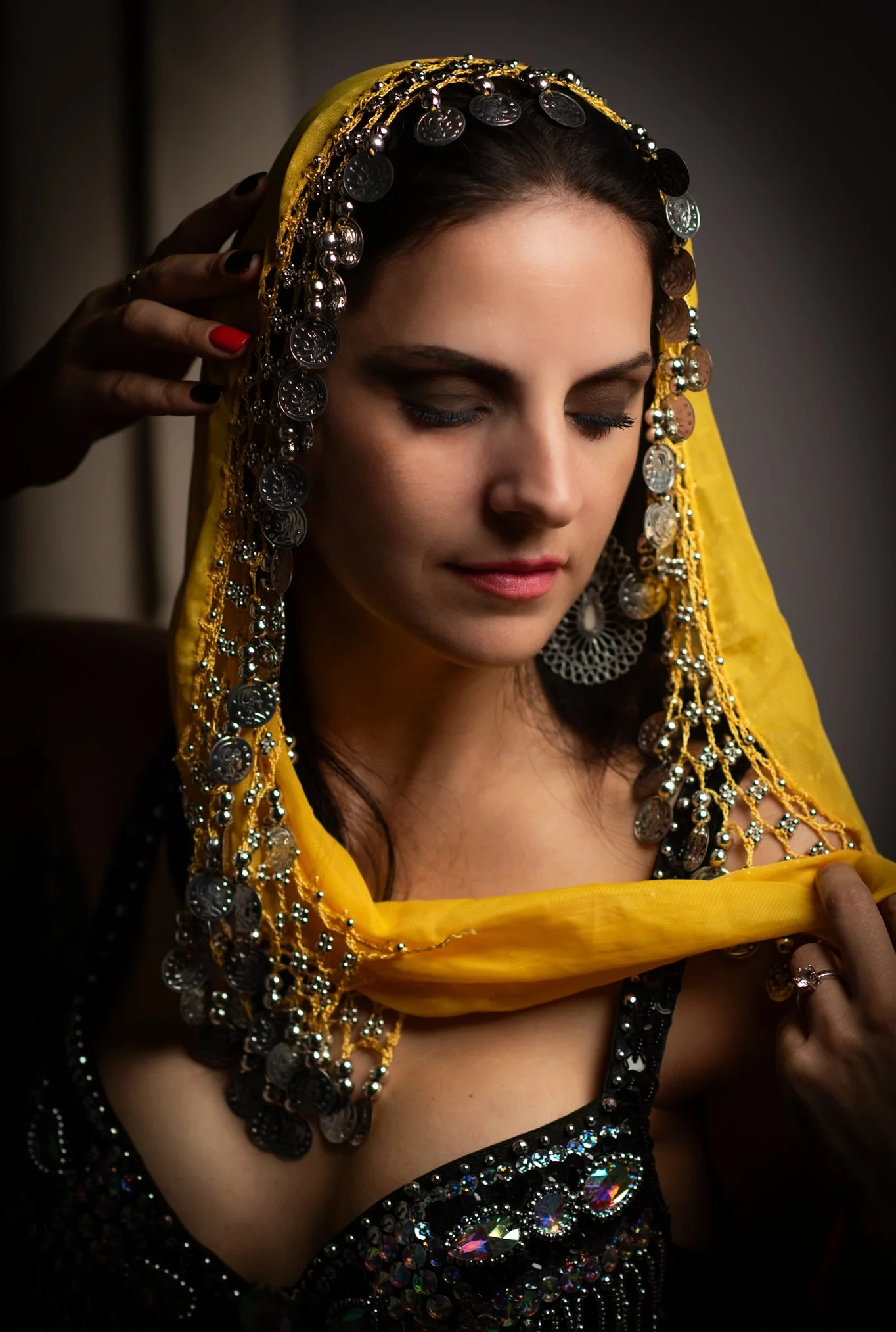 A woman with closed eyes wearing a yellow headscarf and elaborate jewelry, including earrings and necklaces with coins and beads, being assisted by a hand with red nail polish.