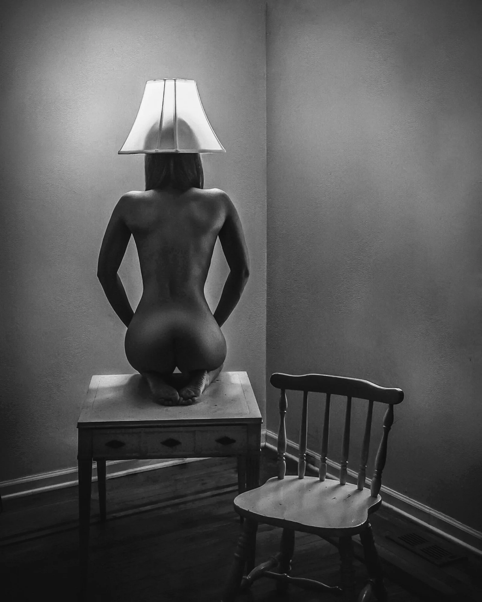 A black and white photo of a woman kneeling on a table with her back turned, placing a lamp on her head, in a room with empty chairs and plain walls.
