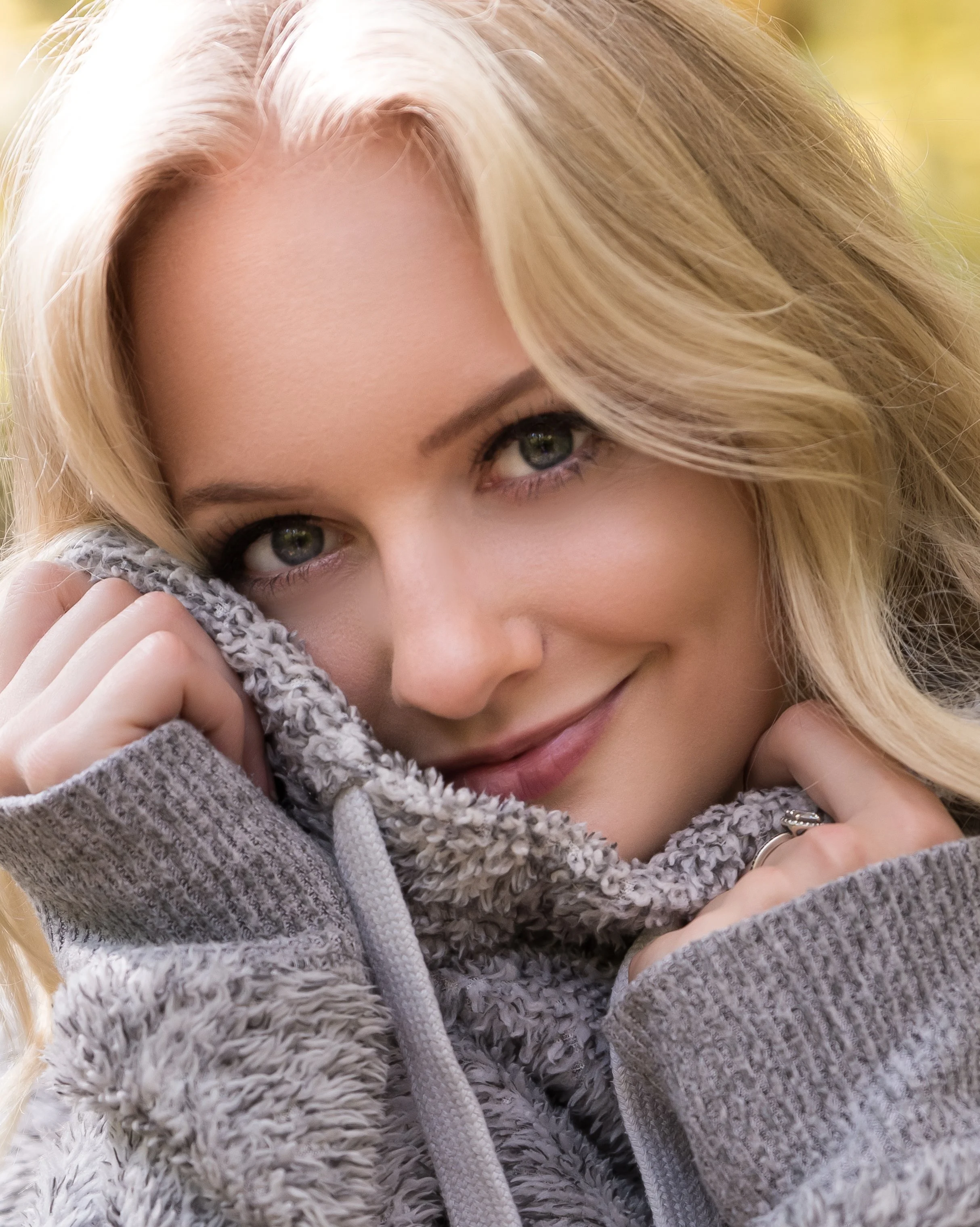 Close-up of a woman with blonde hair, green eyes, and a slight smile, wearing a gray, fuzzy hoodie, resting her face on her hand, outdoors with blurry background.