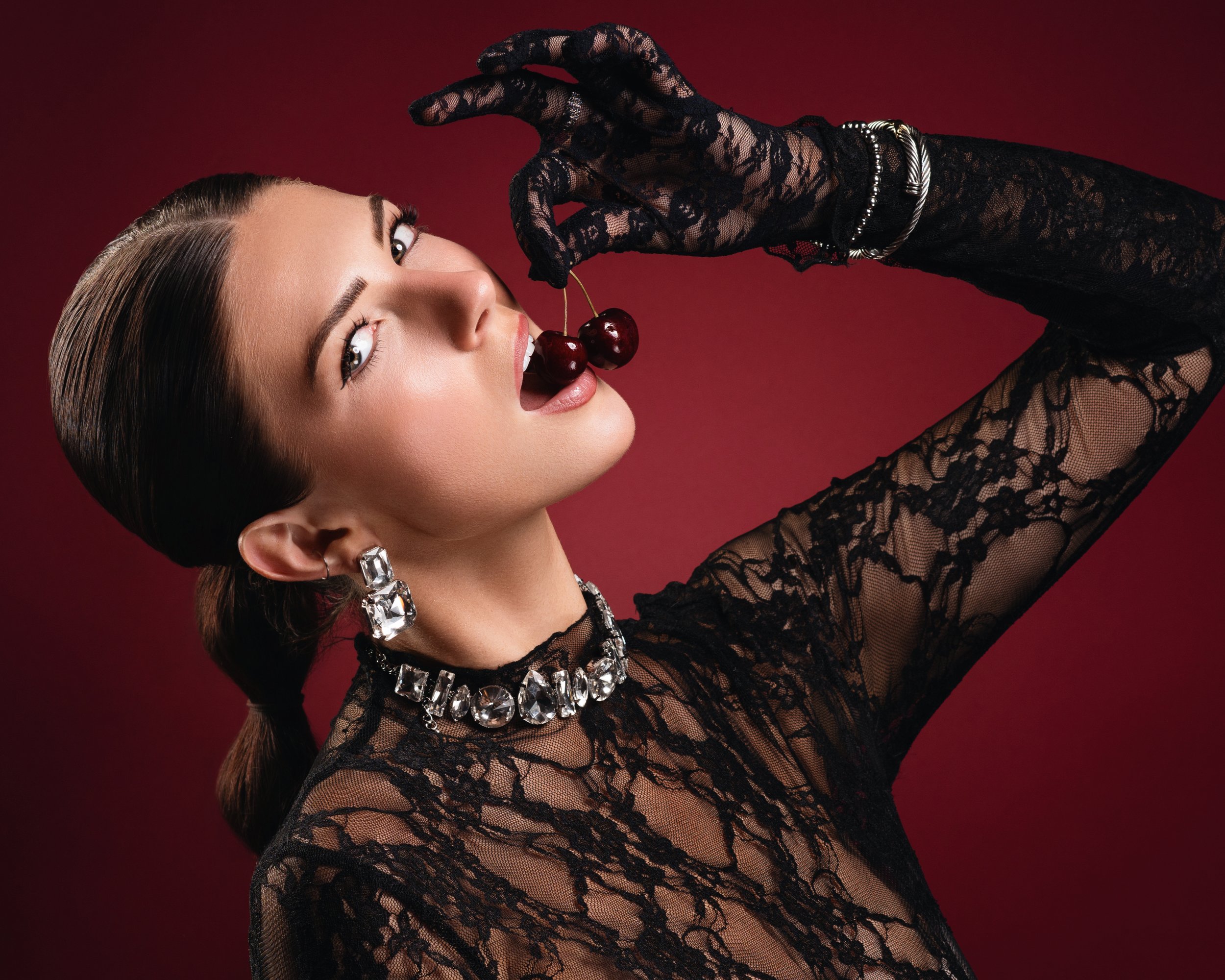 A woman with slicked-back hair and makeup biting a cherry in front of a red background, wearing lace clothing, large gemstone necklace, and earrings.