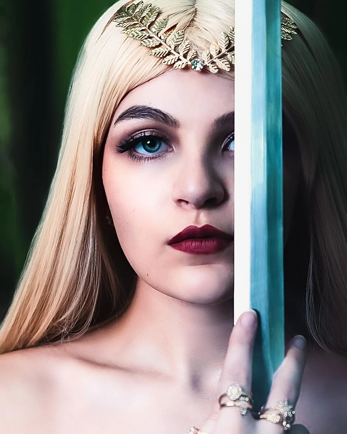 Close-up of a young woman with long blonde hair, wearing a gold leaf crown with a gem, holding a mirror partially covering her face, with striking blue eyes and dark red lipstick.