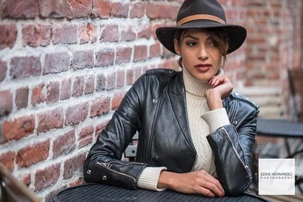 A woman sitting at an outdoor table against a brick wall, wearing a black leather jacket, cream turtleneck sweater, and a black hat with a tan band.