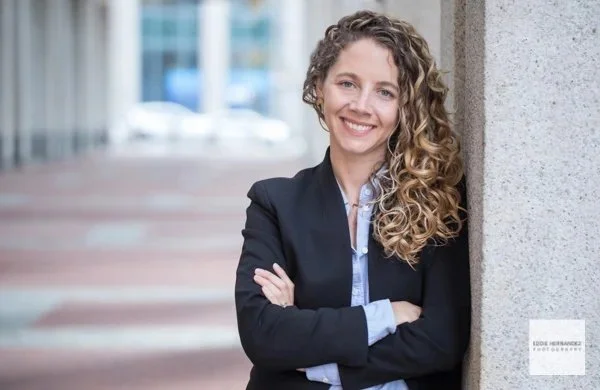 Smiling woman with curly hair leaning against a concrete pillar in an urban setting, wearing a dark blazer and light blue shirt.