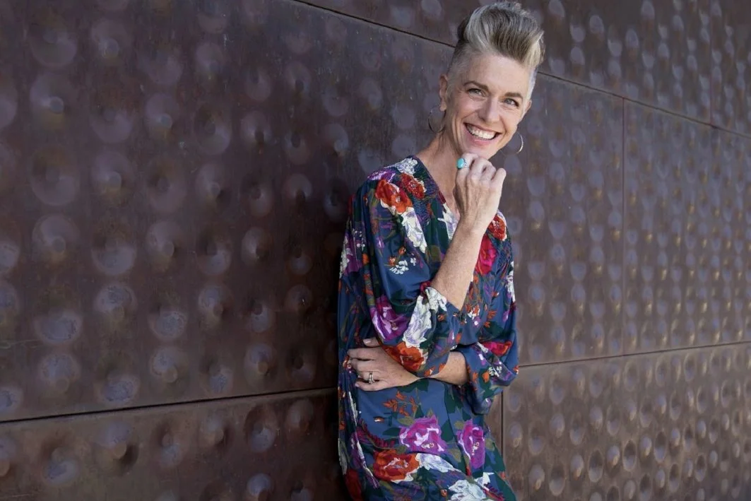 A woman with short blonde hair, wearing a colorful floral dress, leaning against a textured dark wall, smiling and resting her chin on her hand.