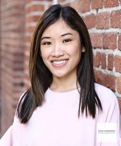 Young woman with long dark hair and light skin smiling, standing in front of a brick wall, wearing a white top.