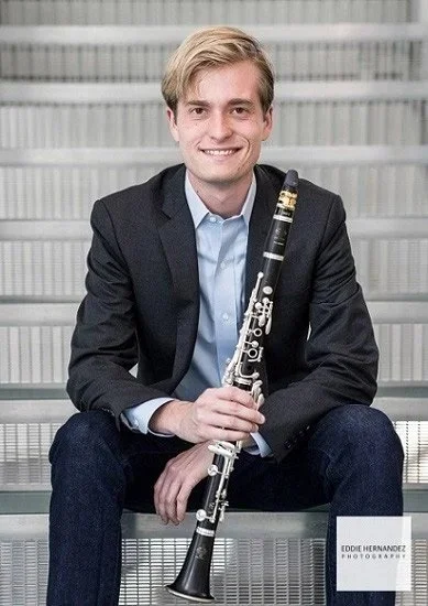 Young man in a dark blazer and light blue shirt, sitting on metal stairs, holding a clarinet. Background shows a modern building with horizontal blinds.