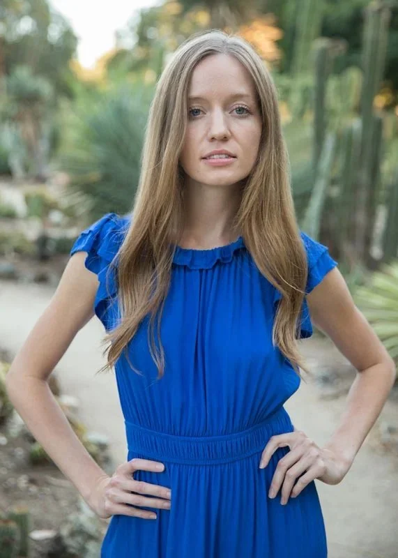 A young woman with long, wavy blonde hair wearing a royal blue dress with ruffled sleeves and standing outdoors with greenery in the background.