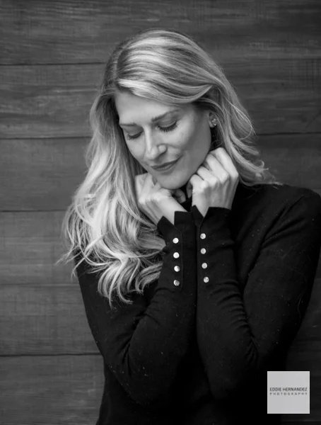 A woman with long, wavy blonde hair wearing a black sweater, standing in front of a wooden wall, with her eyes closed and her hands gently touching her neck.