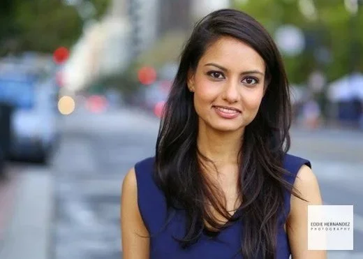 A young woman with long dark hair wearing a blue sleeveless top standing outdoors on a city street.