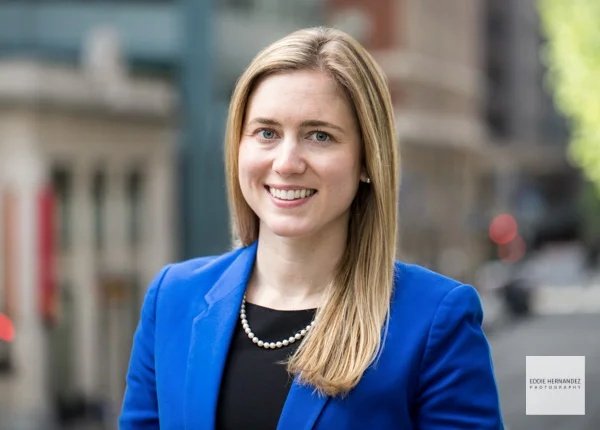 A woman with long blonde hair wearing a blue blazer and pearl necklace, smiling outdoors in an urban setting.