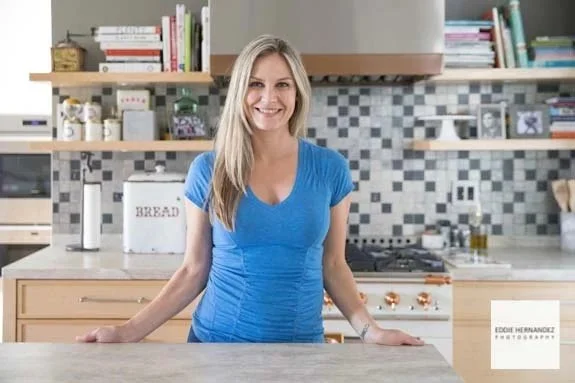 A woman with long blonde hair smiling in a modern kitchen, wearing a blue shirt, with shelves of books and kitchen items in the background.