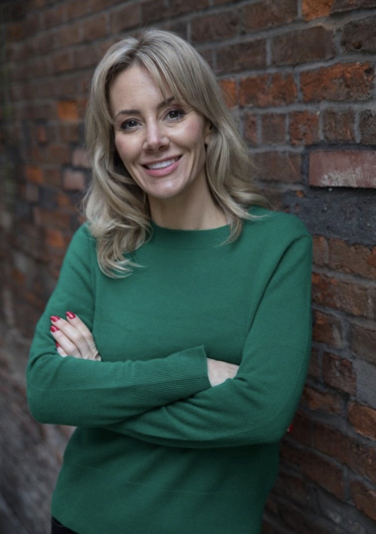 A smiling blonde woman with wavy hair, wearing a green sweater and red nail polish, standing against a brick wall with crossed arms.
