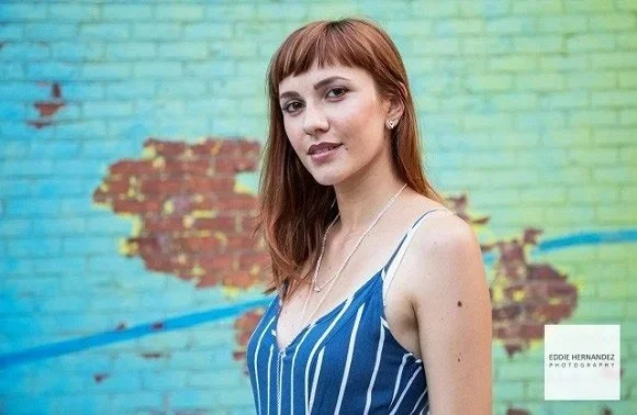A young woman with glasses and reddish-brown hair stands in front of a colorful brick wall with peeling paint, wearing a blue and white striped sleeveless top.