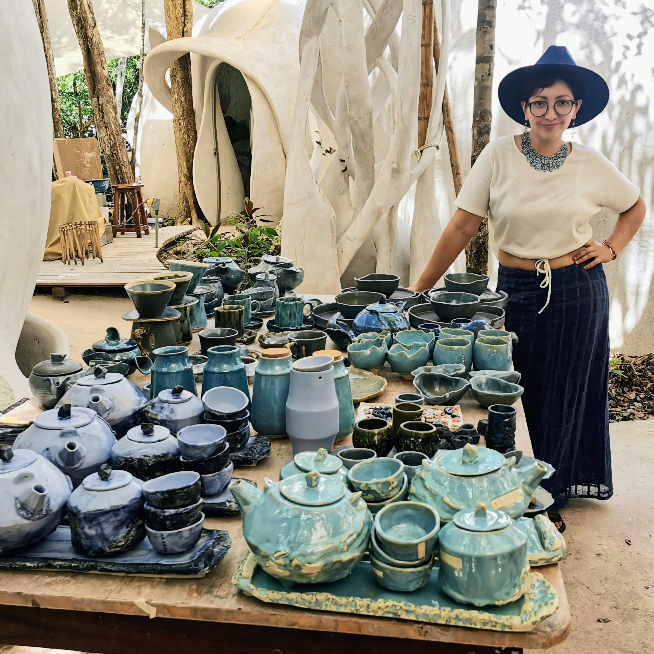 Table filled with assorted handmade ceramic pottery, including bowls and cups, at an outdoor craft market with a person standing beside it, wearing glasses, a wide-brimmed hat, and a white top. Mexican potter.