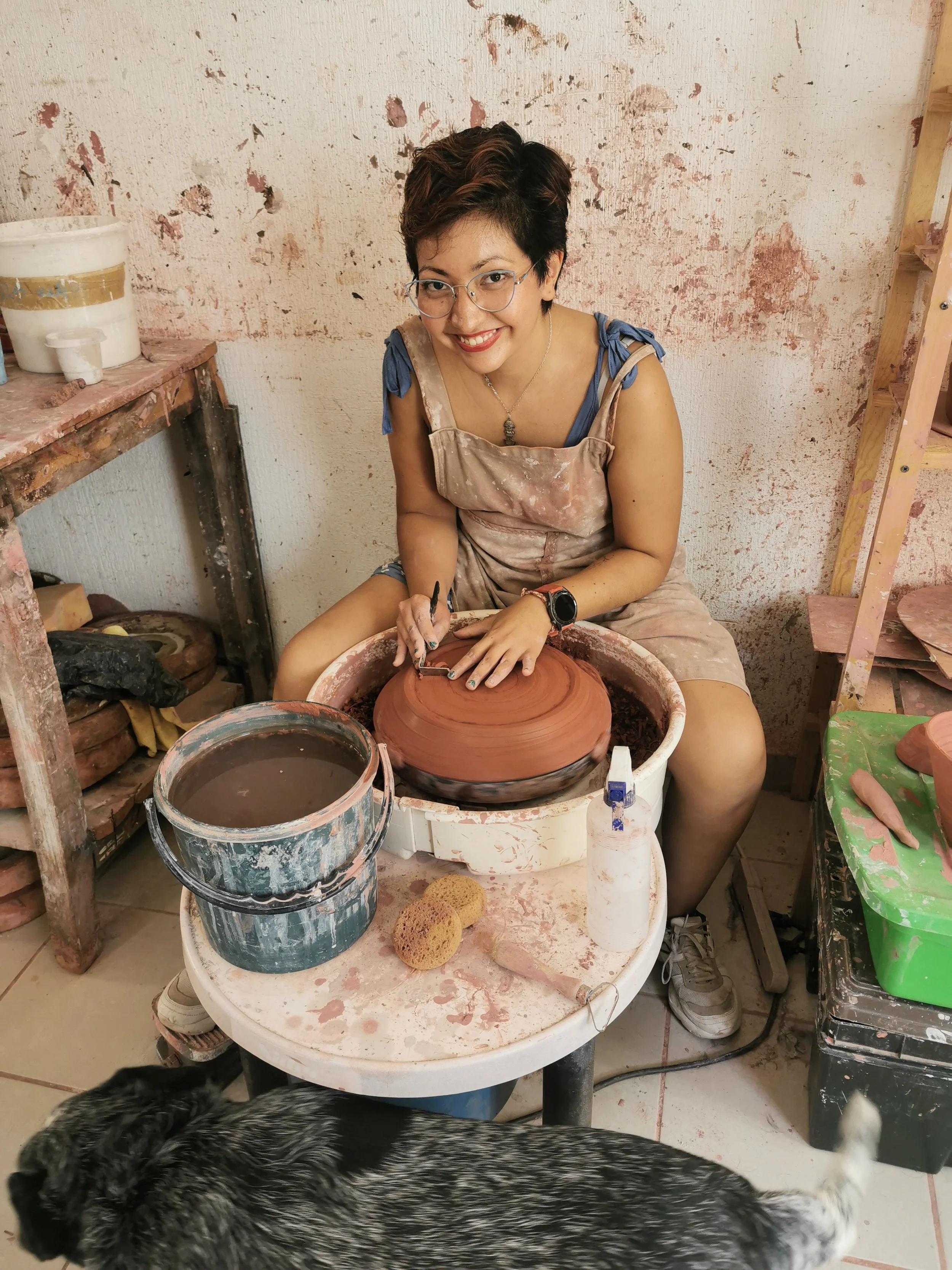 A woman with glasses and short brown hair, wearing a beige apron and blue shirt, is sitting at a pottery wheel shaping a clay piece. The background is a textured wall with paint splatters. There are buckets, tools, and a small green table with pottery supplies around her.