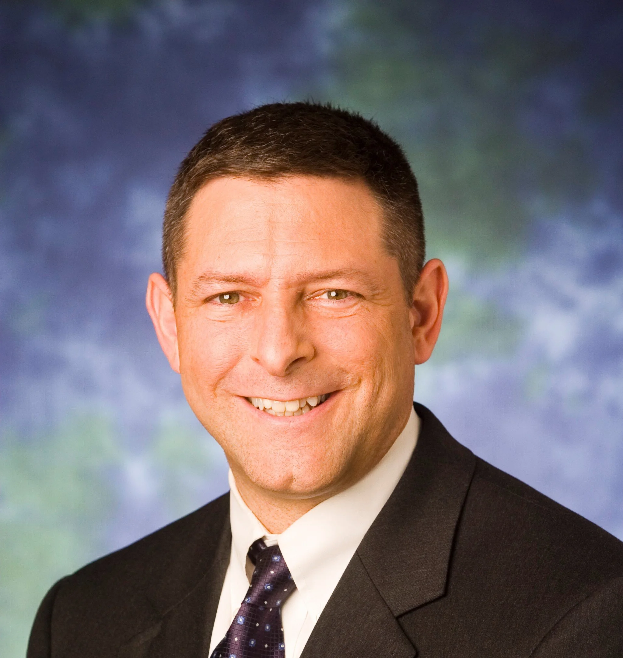 Portrait of a smiling man with short dark hair, wearing a black suit, white shirt, and a dark purple tie with a shiny pattern.