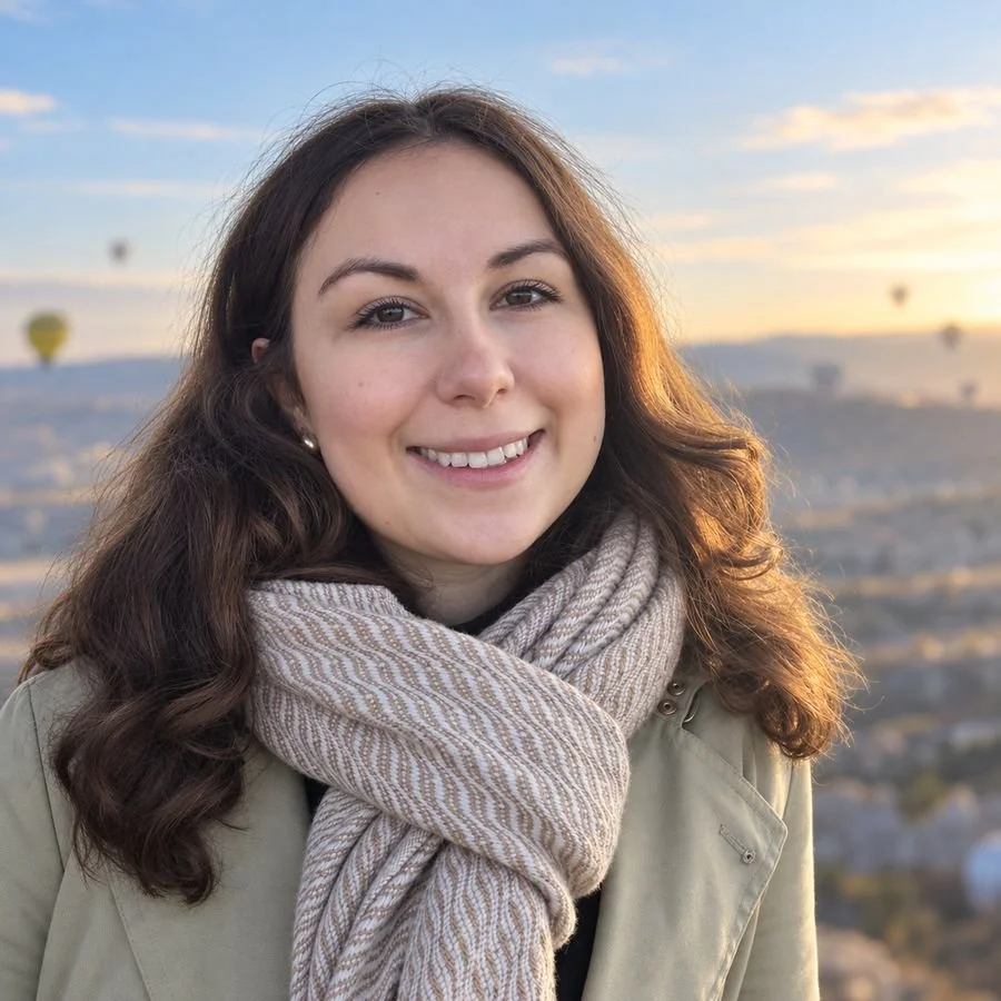 Young woman with brown wavy hair smiling outdoors during sunset, wearing a beige scarf and coat, with hot air balloons in the sky behind her.