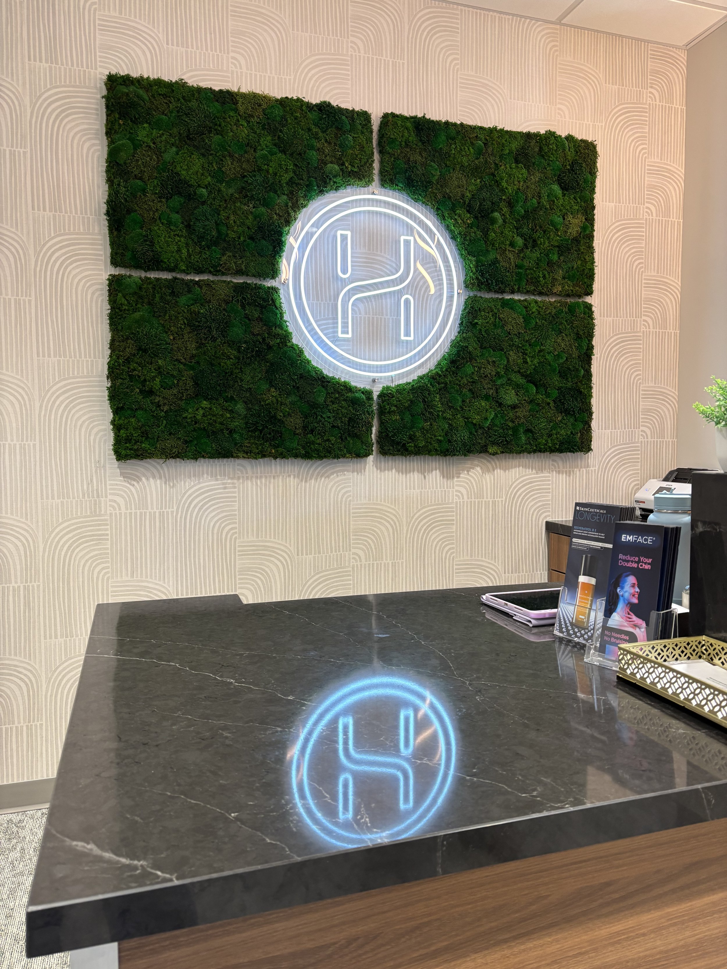 A black marble countertop with a glowing blue and white circular logo reflection on its surface. In the background, there is a green moss wall with a partial illuminated logo, a beige patterned wall, and a reception desk with brochures and a tablet.