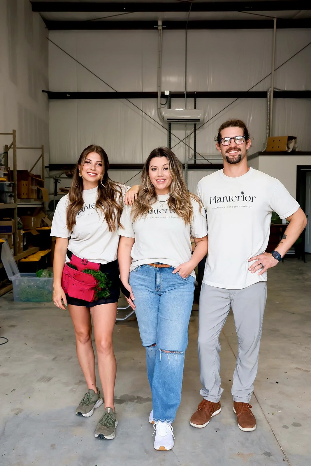 Three young adults, two women and one man, standing together in a warehouse-like space, smiling at the camera. All are wearing white t-shirts with the word 'Planterior' printed on them.