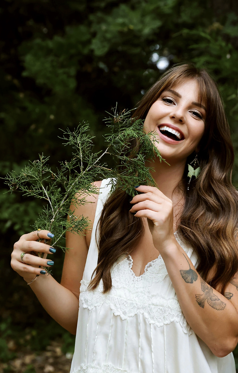 A woman with long brown hair, wearing a white lace dress, smiling and holding a green leafy branch in an outdoor wooded setting.