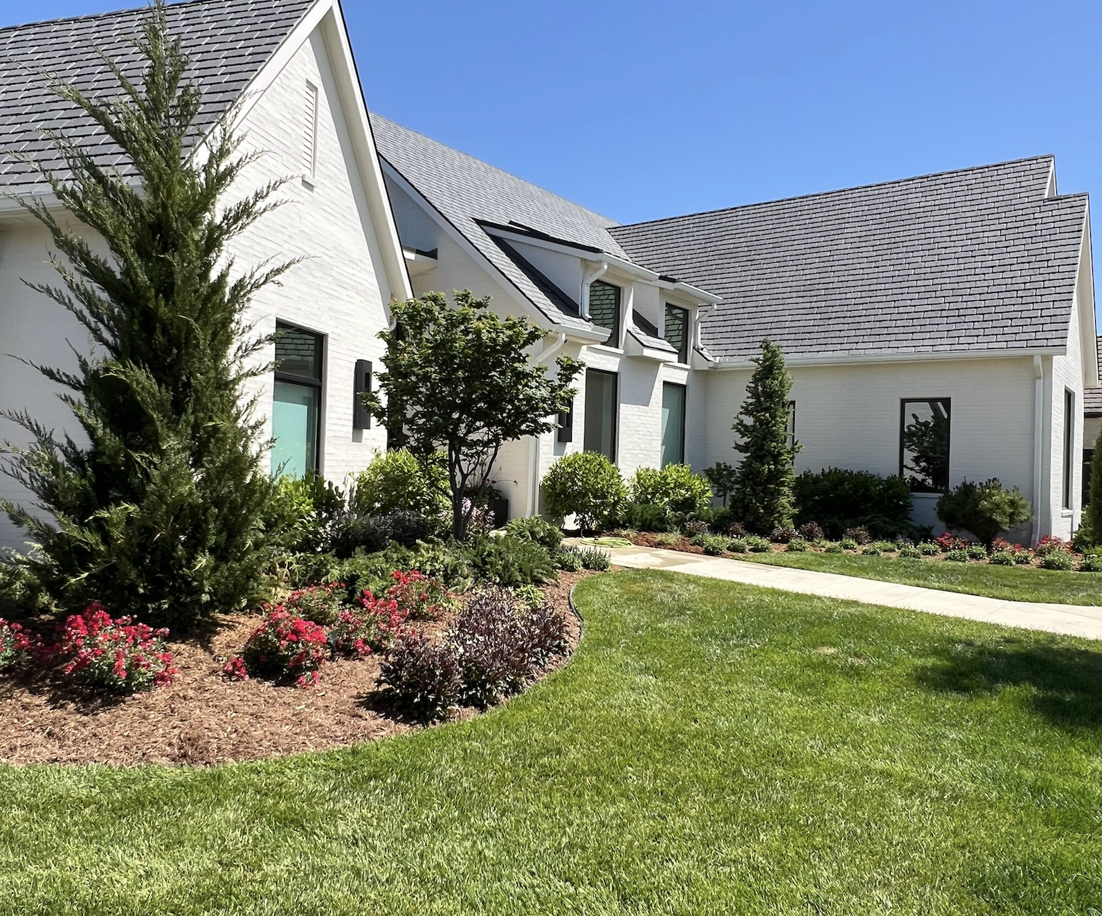 Front view of a modern white house with a landscaped yard, green trees, and colorful flowers under a clear blue sky.
