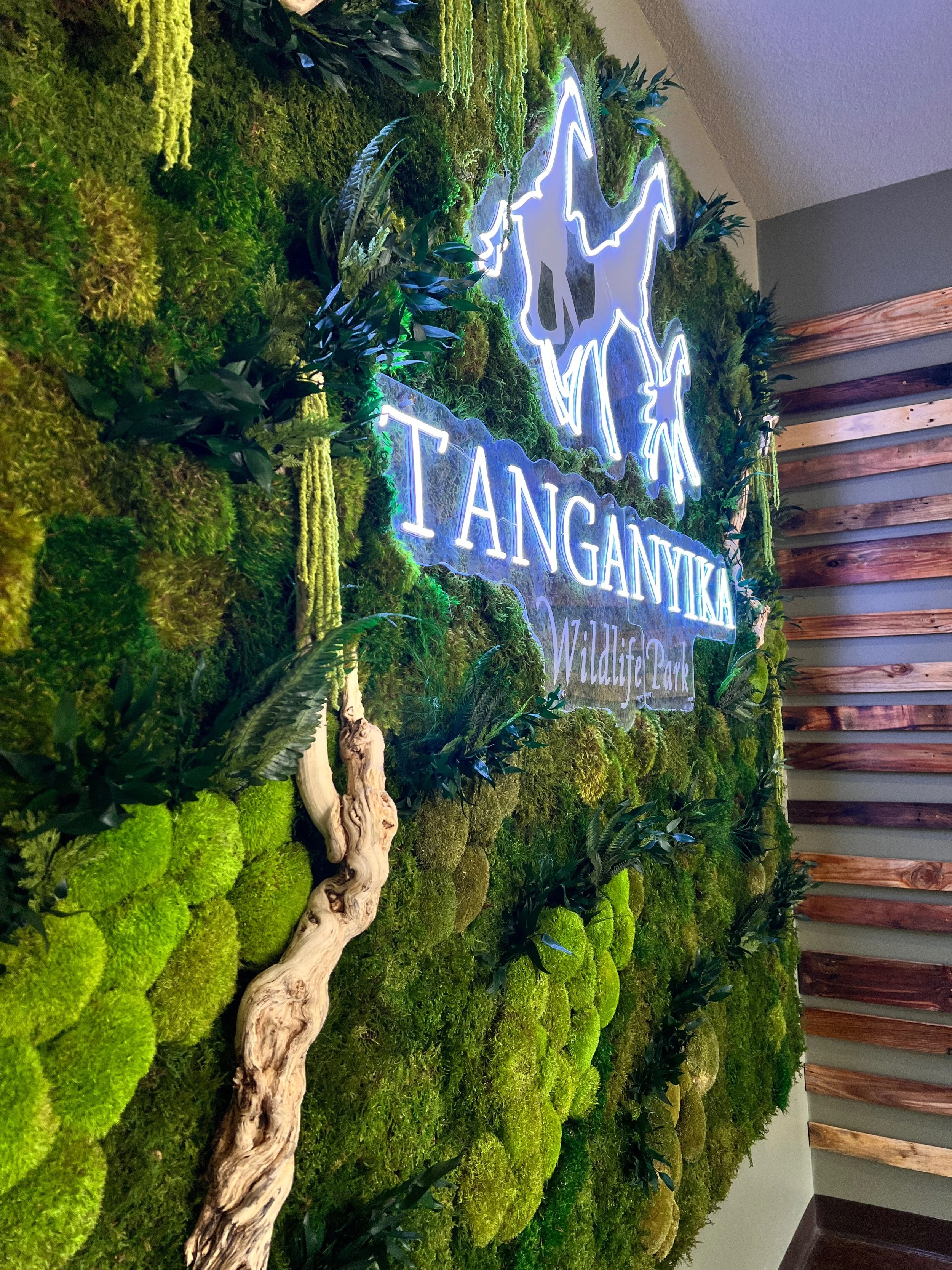 A wall covered with lush green moss and plants, with a neon sign of a stylized fox and the words "TANGANYIKA Wildlife Park".