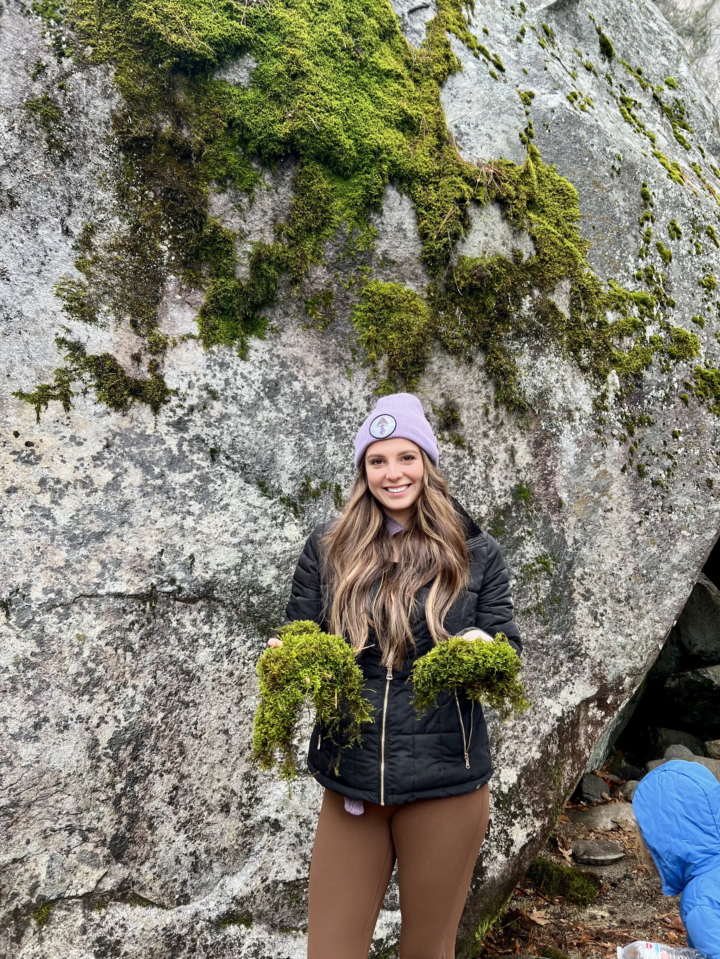 A smiling woman holding two small round moss-covered objects, standing in front of a large moss-covered rock with a person dressed in blue partially visible at the bottom right.