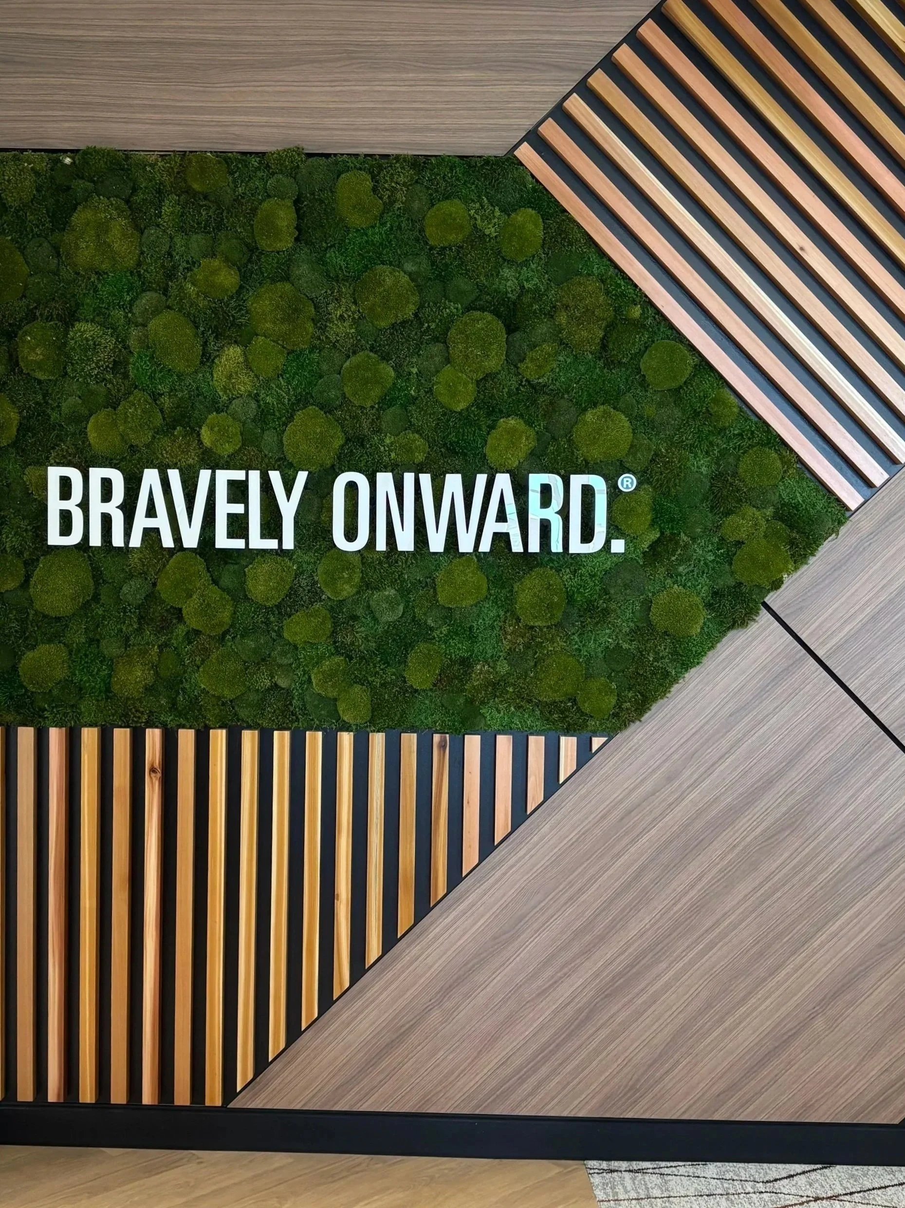 Interior wall decor with wooden slats, green moss or plant wall, and a quote 'Bravely Onward' in white text.