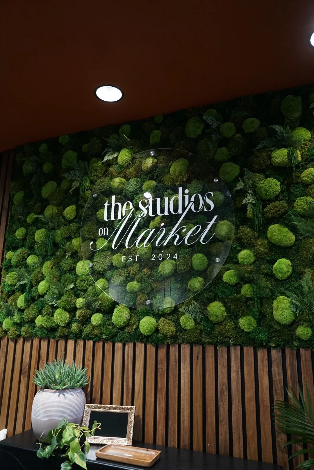Interior wall with green moss decor, a round glass sign that reads 'the studios on Market EST. 2024', and a table with potted plants and picture frames.