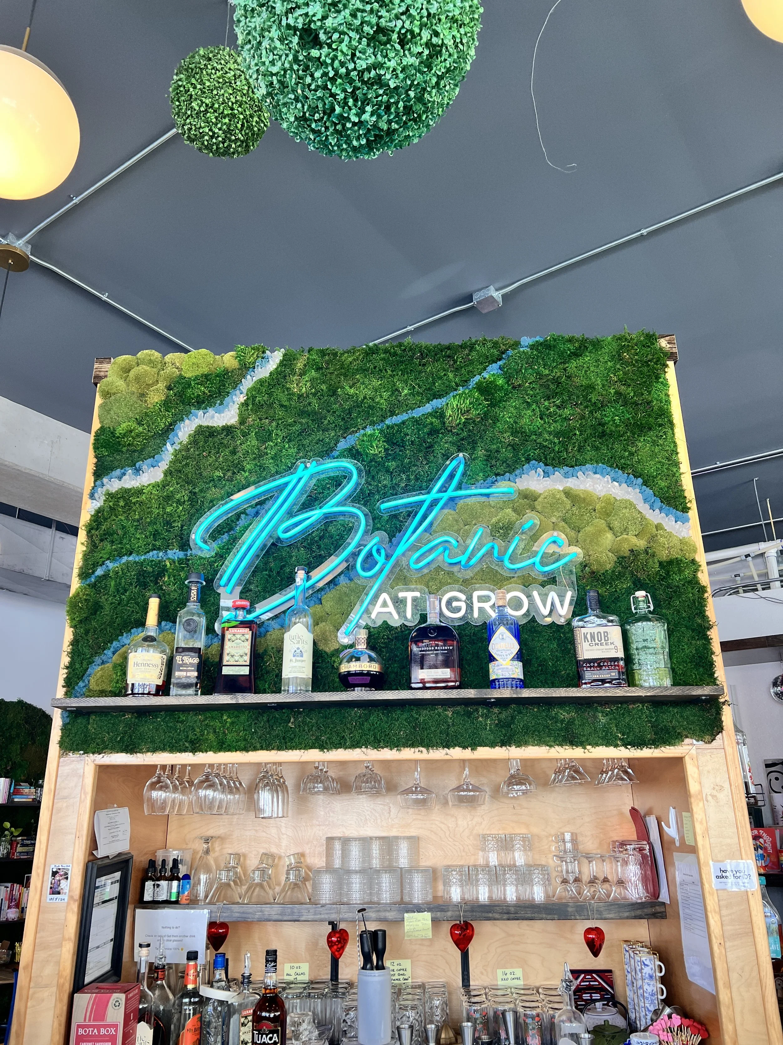 Wooden bar shelf with various liquor bottles, glasses, and bar supplies, decorated with a green wall and neon sign reading 'Botanic At Grow' above the bar area.