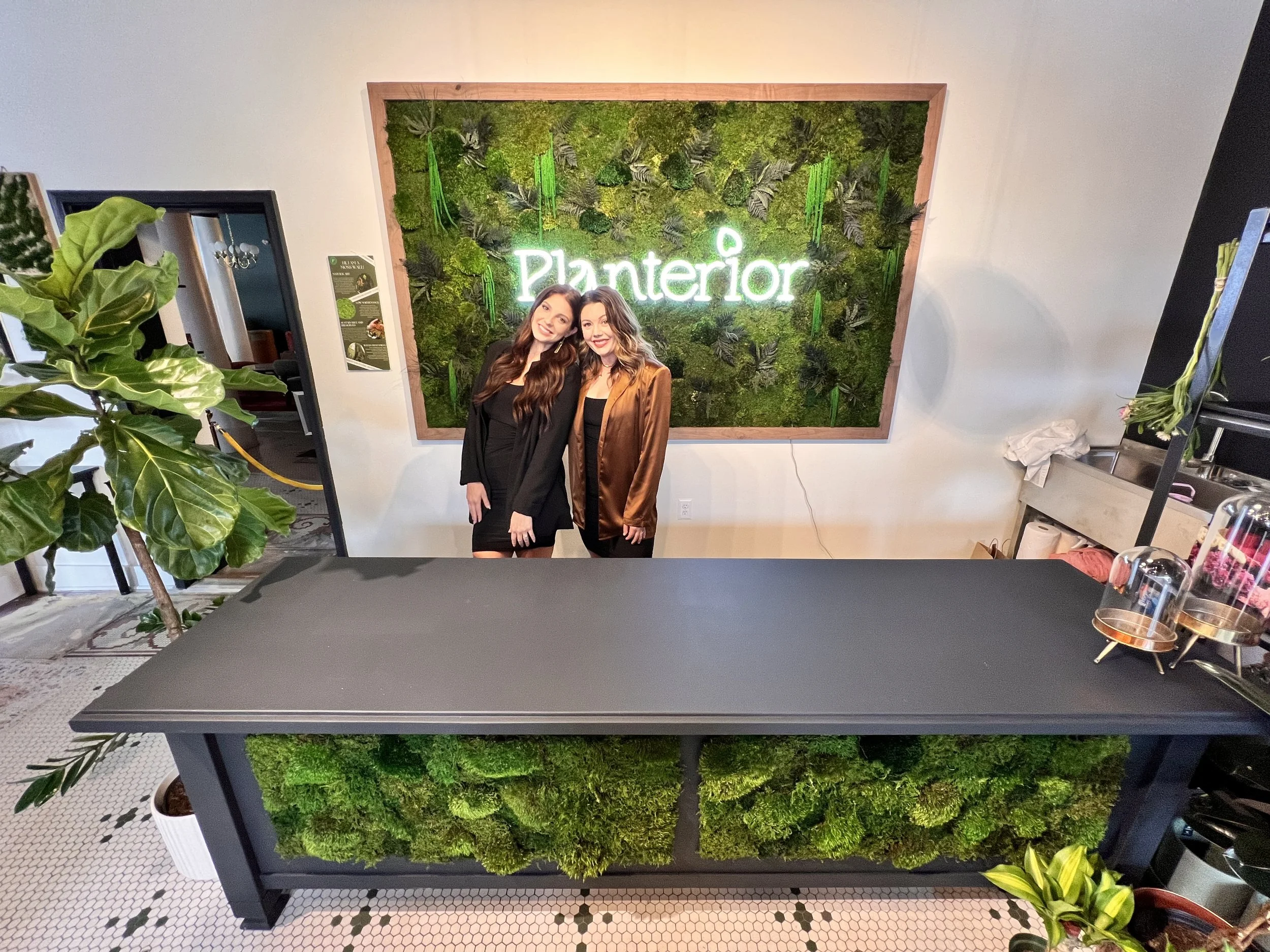 Two women standing in front of a green moss wall with the word 'Planterior' illuminated in white lights.