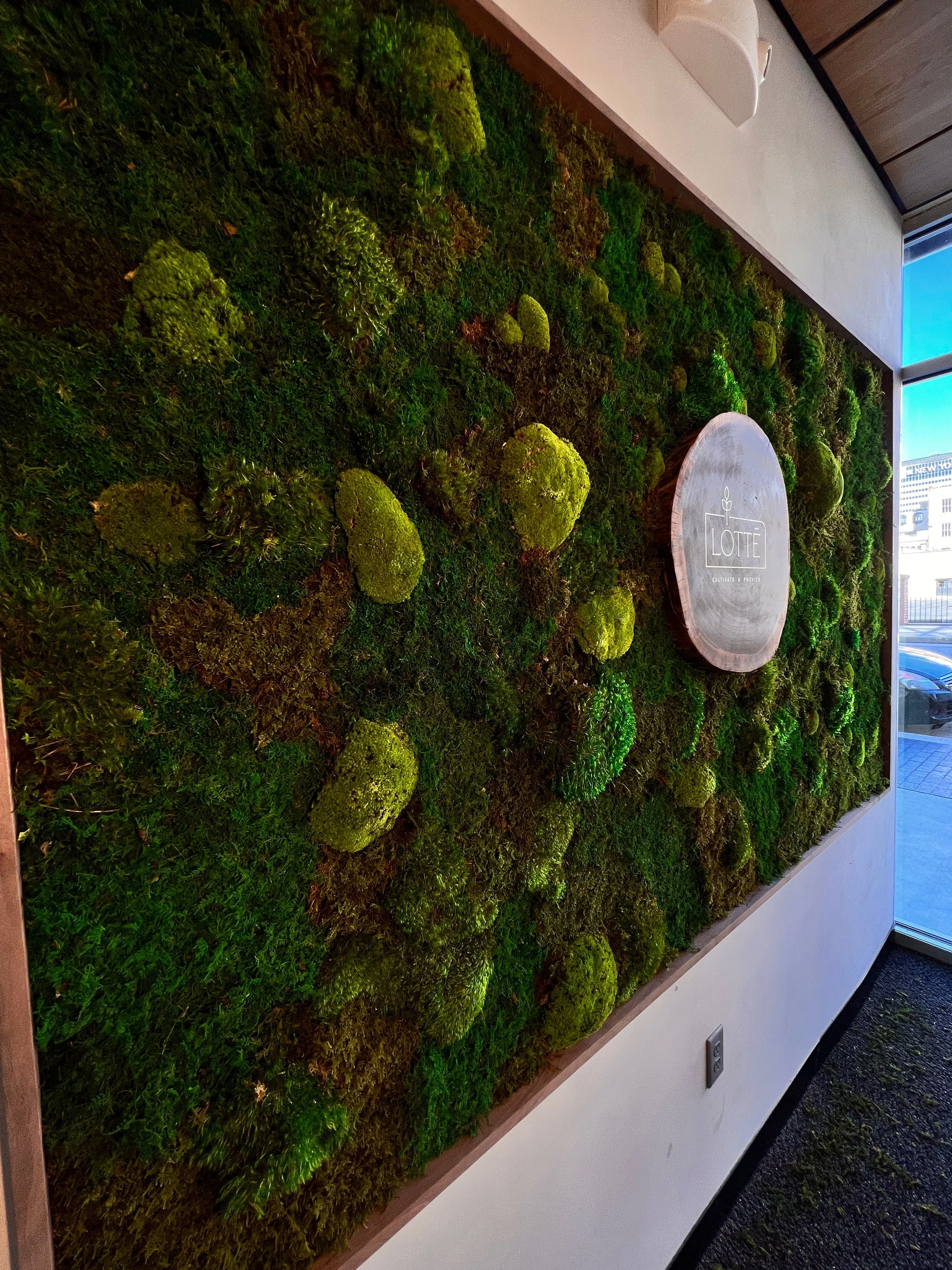 Interior wall decorated with lush green moss and plants, featuring a circular wooden sign with the word 'LOTTE' and an outline of a plant, in a modern building with large windows showing a cityscape outside.
