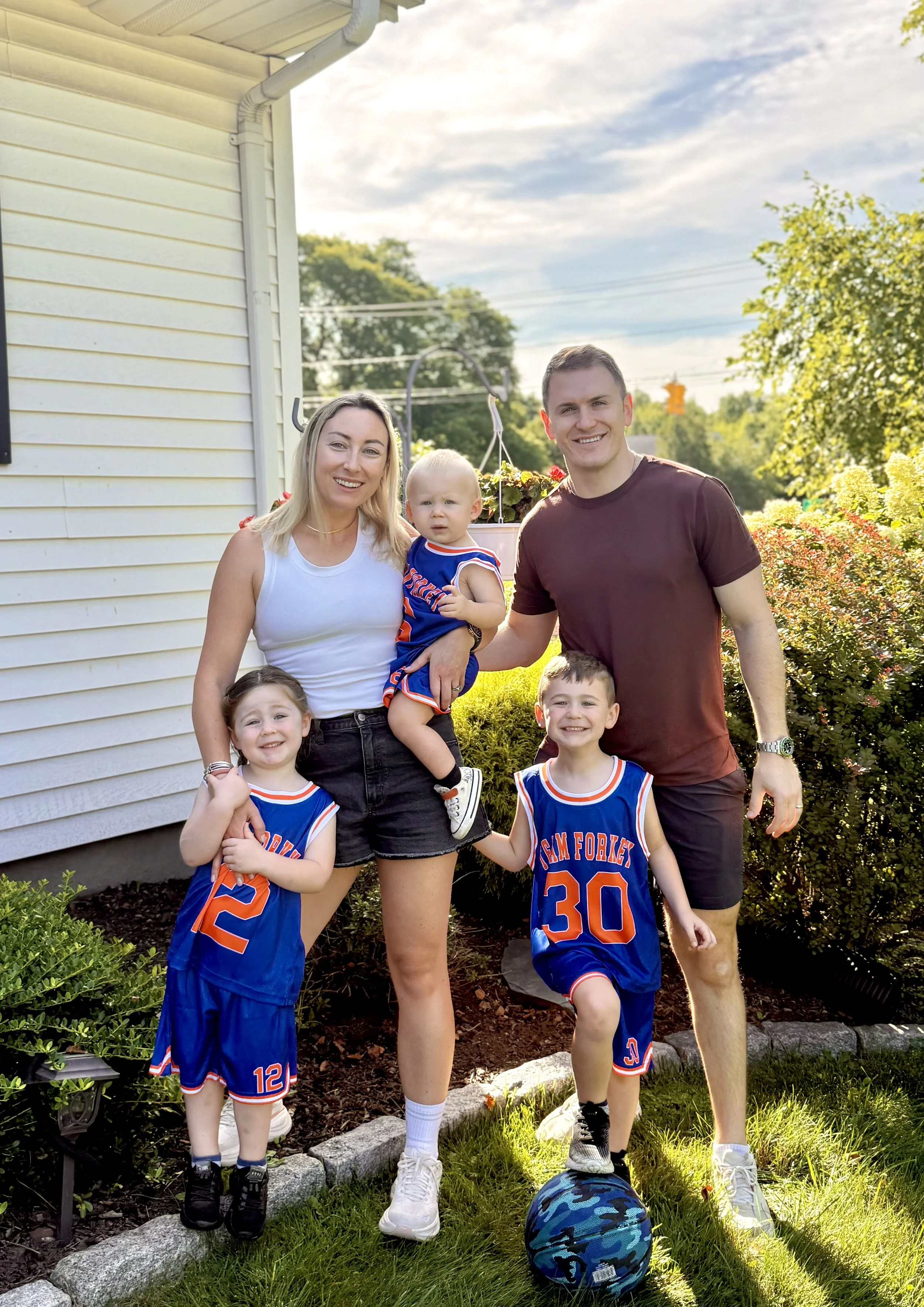 A family outdoor photo with three adult and three children. Two children in basketball uniforms and the adults, a woman and a man, standing behind them, on a garden border with lush greenery. The children and adults are smiling on a sunny day.