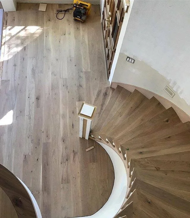 Interior view of a spiral staircase with wooden steps and a wooden floor, partially painted white wall, with a small table, a power tool, and a wooden panel leaning against the wall.